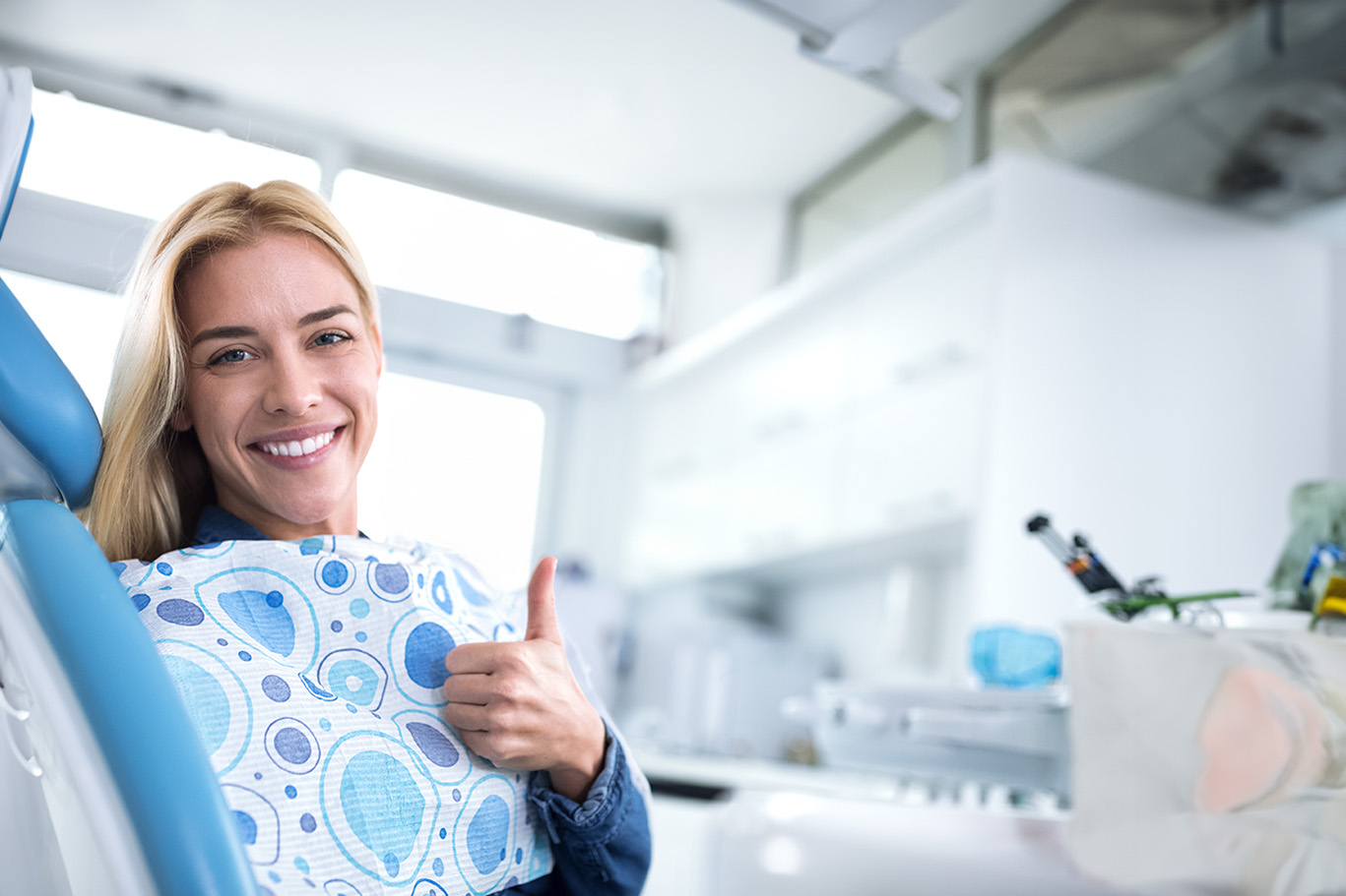 A woman in a dental office, smiling and giving a thumbs-up while wearing a blue apron with white polka dots.